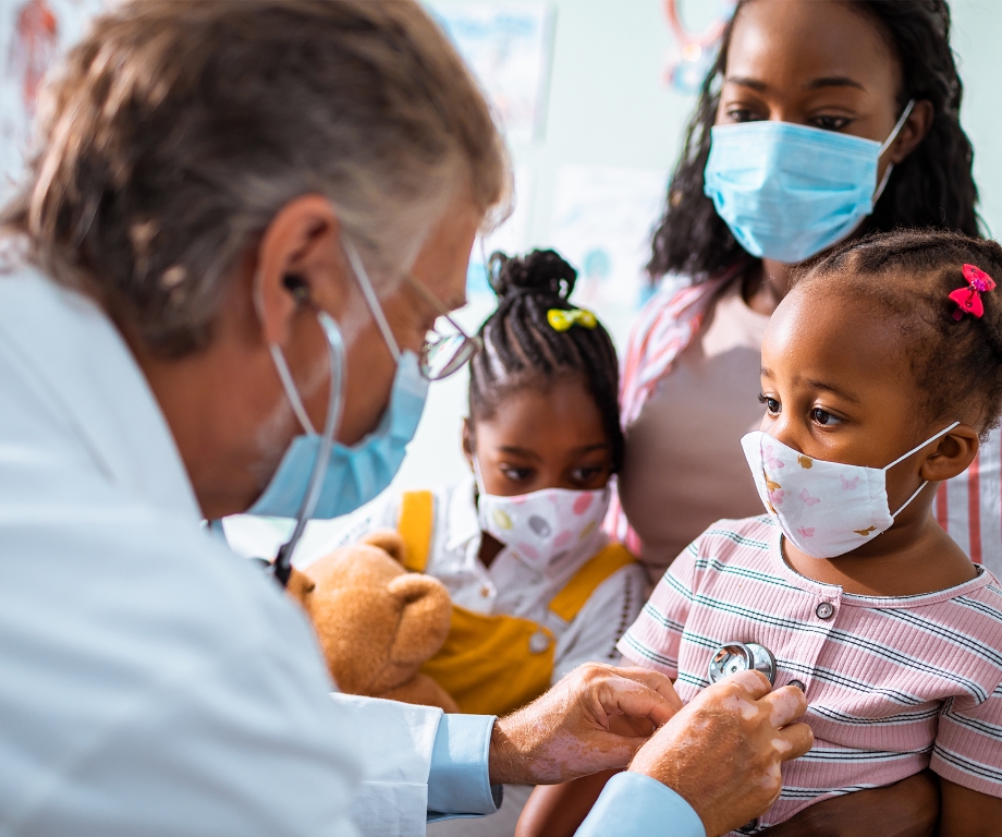 A doctor listens to a girl's heart while her mother holds her and her sister.